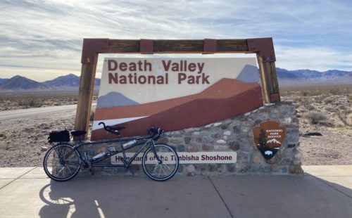 A gray tandem bike in front of a National Park Service sign for Death Valley National Park, the Homeland of the Timbisha Shoshone. The desert sprawls towards mountains in the background.
