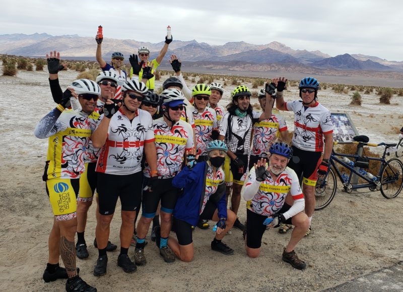 15 stokers and guides gather for a group photo wearing our Blind Stokers Club shirts, bike shorts, and helmets. Many of the club members have on sunglasses, and we are standing off the road in the sand and dirt with mountains in the background. 