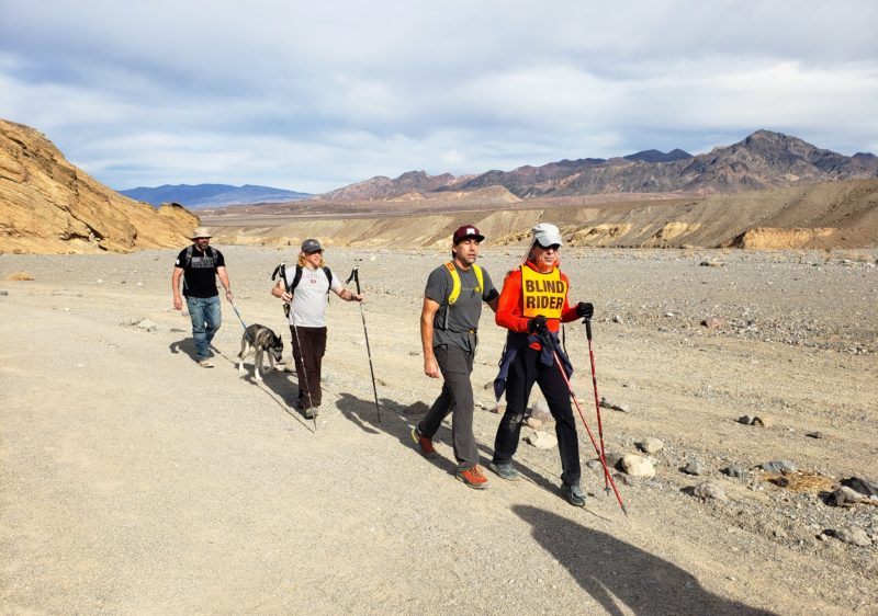 Erik links arms with a BSC member wearing a “Blind Rider” while hiking in the desert; two (2) more BSC members walk behind them, one with poles, another with a husky dog.