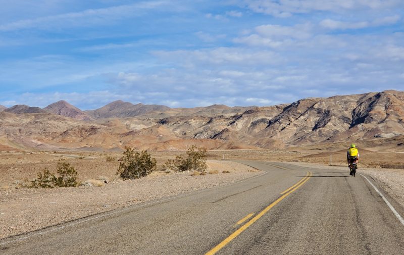 A BSC stoker and captain ride off down a paved road that cuts between low-lying mountains in Death Valley National Park. 