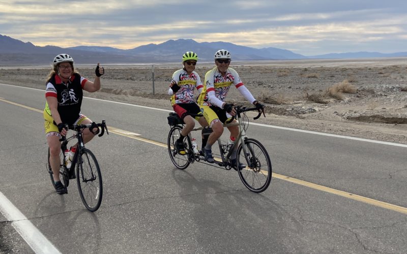 A stoker and captain riding a tandem bike in Death Valley – kitted out in their BSC gear – alongside a single, sighted rider.
