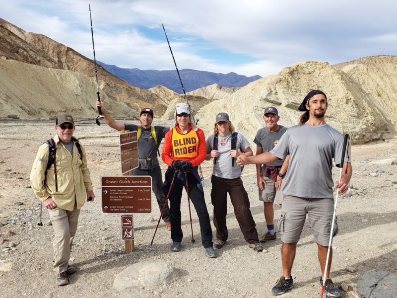Erik, Mark, Bob, and two (2) other BSC members taking a group photo at the Gower Gulch Junction in Death Valley National Park. Mark is in the foreground wearing all gray and holding a white and red cane. 