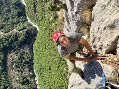 Erik climbing the Verdon Gorge. He is wearing a red Camp USA climbing helmet, grey t-shirt and climbing pants, and is harnessed and roped up. He is looking at the camera with the Verdon River snaking hundreds of feet below him through the heart of the valley.