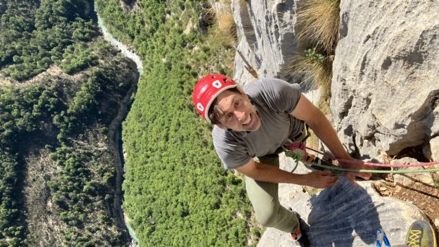 Erik climbing the Verdon Gorge. He is wearing a red Camp USA climbing helmet, grey t-shirt and climbing pants, and is harnessed and roped up. He is looking at the camera with the Verdon River snaking hundreds of feet below him through the heart of the valley.
