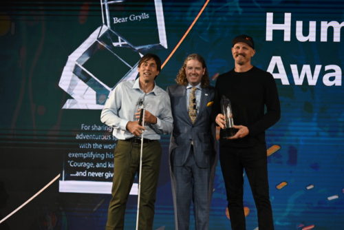 Erik and Mick receiving and posing with their award, with Dr. Mark Lyons between them. Mick is wearing a black cap, shirt, and pants. Erik is in a light colored button up and dark pants. Mark is in a blue suit and gold tie.