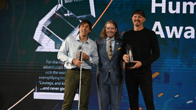 Erik and Mick receiving and posing with their award, with Dr. Mark Lyons between them. Mick is wearing a black cap, shirt, and pants. Erik is in a light colored button up and dark pants. Mark is in a blue suit and gold tie.