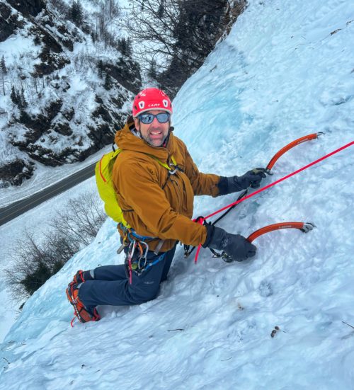 Erik midway up an ice climb with his axes and crampons holding him to the surface as he looks over his right shoulder toward the camera.