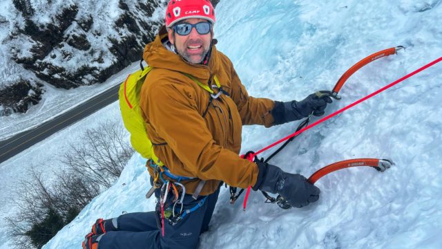 Erik midway up an ice climb with his axes and crampons holding him to the surface as he looks over his right shoulder toward the camera.